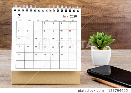 July 2026 desk calendar on wooden table with black smartphone. 128794425