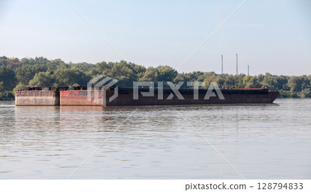 Two rusting barges float on a serene river surrounded by a lush forest 128794833