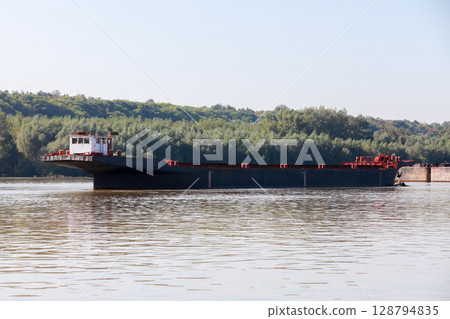 A large cargo ferry navigating calm waters of a river with a forested background A large cargo ferry navigating calm waters of a river with a forested background 128794835
