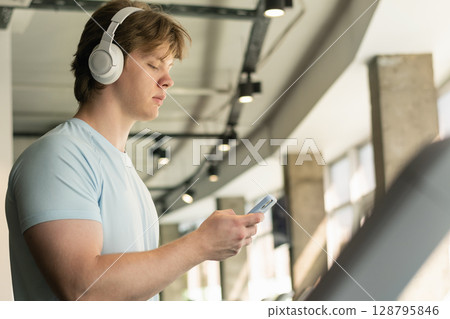 Athlete man with cellphone or mobile phone running track on treadmill at gym. Warming up. Preparation for cardio training. Work out at fitness center. Close up. 128795846