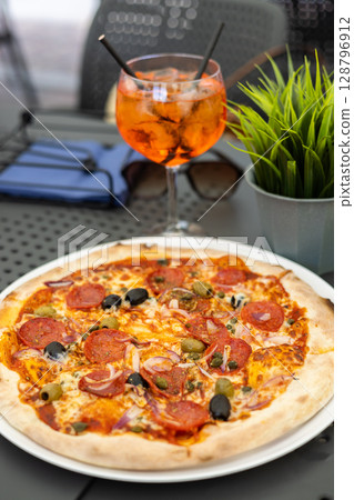 Italian delicious pizza with salami pepperoni and summer Spritz cocktail in a glass with ice and straws stands on a table in a restaurant or cafe 128796912