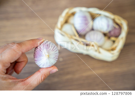 Female hand holding a head of Chinese single clove garlic with a basket full of garlic in the background. Natural food ingredient and kitchen preparation. Female hand holding a head of Chinese single clove garlic with a basket full of garlic in the background. Natural food ingredient and kitchen preparation. 128797806