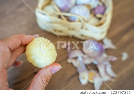 Fingers holding peeled Chinese single clove garlic with basket full of garlic and peel in the background. Cooking preparation, natural seasoning, and fresh ingredient. Fingers holding peeled Chinese single clove garlic with basket full of garlic and peel in the background. Cooking preparation, natural seasoning, and fresh ingredient. 128797855