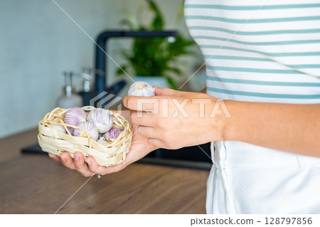 Hands of young woman holding a basket with Chinese single clove garlic on the kitchen table. Organic cooking ingredient, natural food, and culinary preparation. Hands of young woman holding a basket with Chinese single clove garlic on the kitchen table. Organic cooking ingredient, natural food, and culinary preparation. 128797856