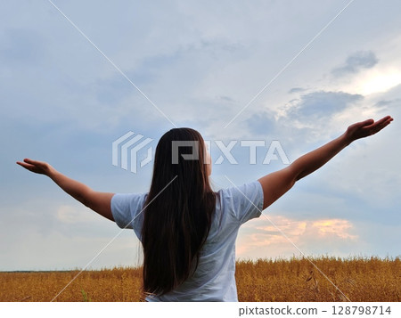 Woman raises her hands to the sky. Young woman stands in middle of wheat field and raised her hands up. Beautiful long hair. Rear view. 128798714