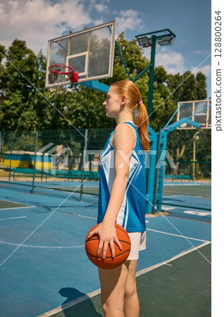 Young woman with basketball standing on outdoor court beneath blue sky 128800264