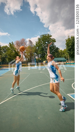 Pair of women engaged in competitive basketball on an outdoor court under a clear sky 128800336