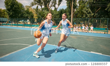 Two women engage in a game of basketball on an outdoor court beneath sunny skies 128800337