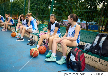 Female basketball athletes sitting on bench in team uniforms at outdoor court 128800345