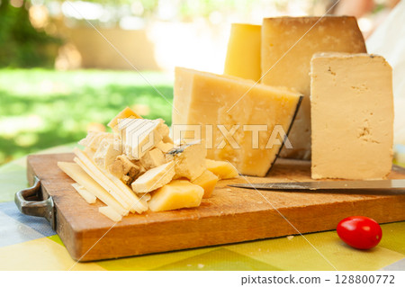 Assorted blocks and slices of hard cheese on wooden board with knife and tomato in outdoor summer setting Assorted blocks and slices of hard cheese on wooden board with knife and tomato in outdoor summer setting 128800772