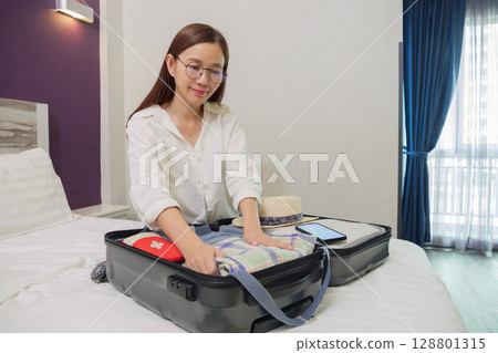 happy Asian woman with smile packing suitcase on bed in bedroom, preparing for summer holidays abroad trip with hat, travel medicine first aid kit, cloth, phone mockup with copy space for advertise 128801315
