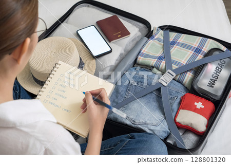 making a check list of things to pack for travel. woman writing checklist item on notebook and packing suitcase to vacation, prepare clothes, medicine, passport into luggage. travel vacation concept 128801320