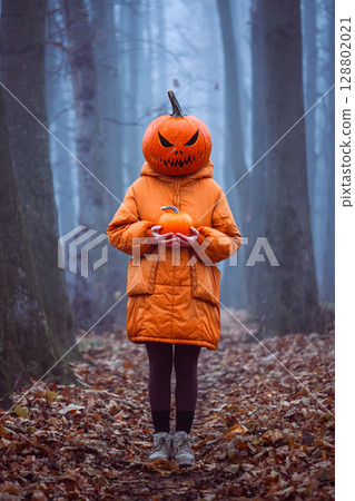 Halloween girl in carved pumpkin mask stands in foggy fall forest 128802021