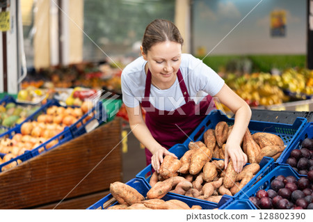 Positive woman seller of fruit and vegetable store work near showcase with garden-stuff, sorting yam Positive woman seller of fruit and vegetable store work near showcase with garden-stuff, sorting yam 128802390