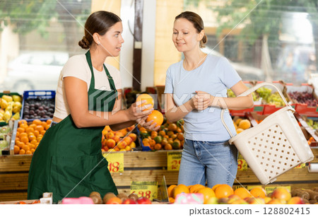 Polite salesgirl offering sweet oranges to female customer in greengrocery store Polite salesgirl offering sweet oranges to female customer in greengrocery store 128802415