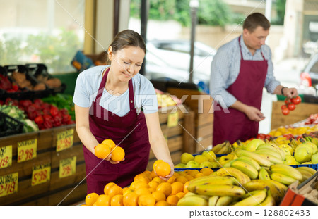 Positive saleswoman in apron offering oranges on supermarket Positive saleswoman in apron offering oranges on supermarket 128802433