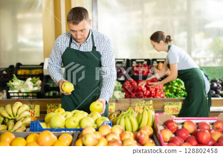 Man seller working in supermarket and lays out apples on counter 128802458