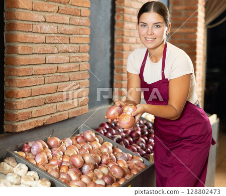 Girl seller working in supermarket and lays out onions on counter Girl seller working in supermarket and lays out onions on counter 128802473