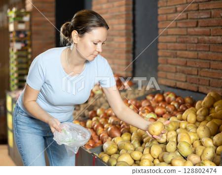 Woman purchaser choosing potatoes at counter in large grocery store Woman purchaser choosing potatoes at counter in large grocery store 128802474
