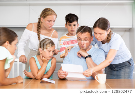Frustrated man sits in kitchen next to family and reads unpleasant news in letter. Frustrated man sits in kitchen next to family and reads unpleasant news in letter. 128802477