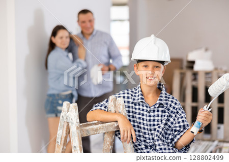 Portrait of builder boy in hard hat with a paint roller. In the background, mom and dad are renovating room Portrait of builder boy in hard hat with a paint roller. In the background, mom and dad are renovating room 128802499