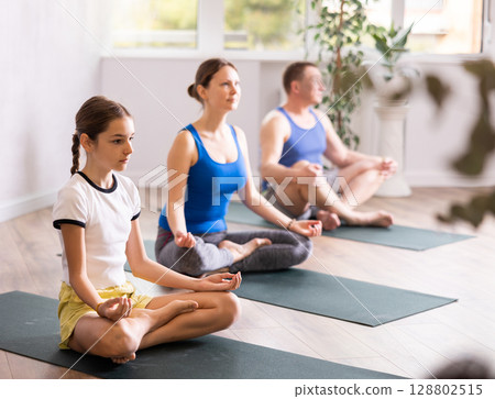 Pre-teen girl performing yoga exercises with brother and parents at gym, family health concept 128802515