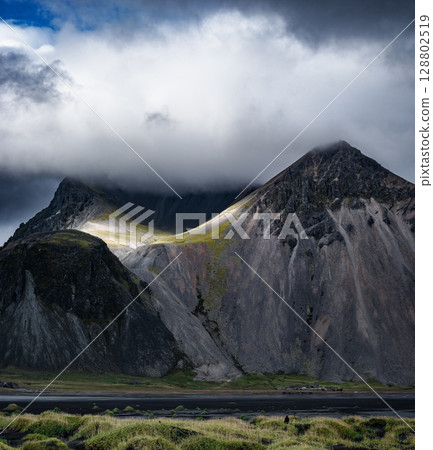 Dramatic view of Vestrahorn mountain in Iceland, partially covered by clouds with soft sunlight on slopes. Concept of isolation, majesty, raw nature, northern silence and geological force. Dramatic view of Vestrahorn mountain in Iceland, partially covered by clouds with soft sunlight on slopes. Concept of isolation, majesty, raw nature, northern silence and geological force. 128802519