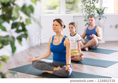 School children and their parents sitting on mats in Lotus pose during family yoga training in gym fitness center indoors 128802538