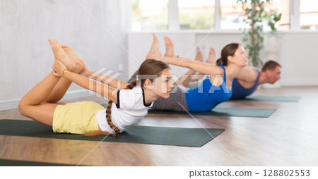 Middle-aged parents and their preteen children practicing Bow Pose or Dhanurasana on mats in studio 128802553
