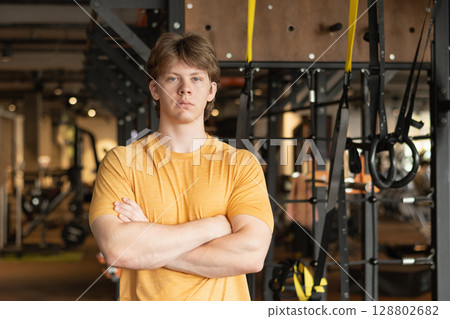 Handsome serious fitness trainer standing wearing orange t-shirt with cross hand at gym. Motivation for regular workout, body building, health and energy. Handsome serious fitness trainer standing wearing orange t-shirt with cross hand at gym. Motivation for regular workout, body building, health and energy. 128802682