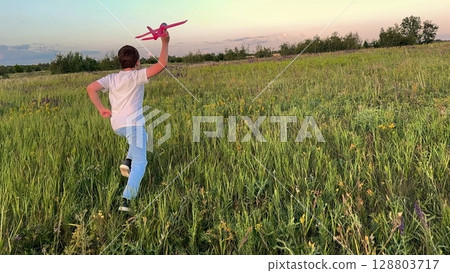 Boy running with toy airplane in a field, Child enjoying outdoor play at sunset, Summer evening fun with a pink glider, Little pilot dreaming big in nature, Carefree childhood moments in green meadow 128803717