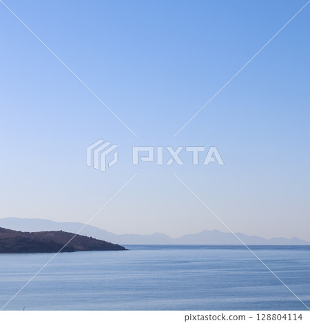 A tranquil blue seascape with a dark coastline in the foreground, and hazy distant mountains fading into a clear, gradient sky. 128804114