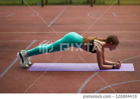 Fit woman performing plank exercise on purple mat outdoors, showcasing strength and determination in a vibrant athletic environment. Workout concept 128804419