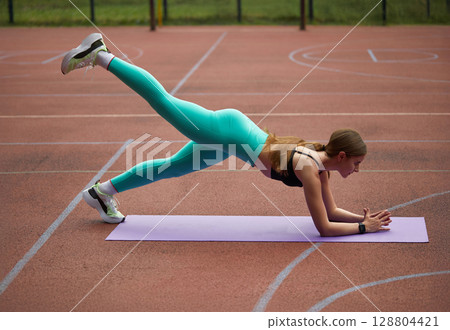 Fit woman exercising outdoors on a yoga mat, performing a plank with leg lift, showcasing strength and determination in a vibrant setting. Workout concept Fit woman exercising outdoors on a yoga mat, performing a plank with leg lift, showcasing strength and determination in a vibrant setting. Workout concept 128804421