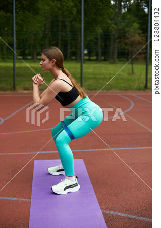 Fit woman performing squats outdoors on a purple mat, showcasing strength and determination in a vibrant green park environment. Workout concept 128804432