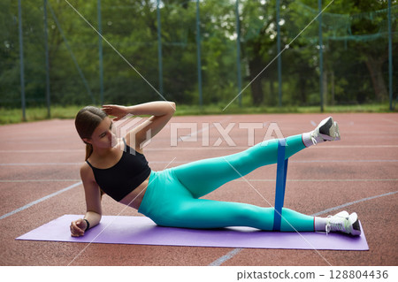 Fit woman exercising outdoors on a yoga mat, performing leg lifts with resistance band, showcasing strength and dedication to fitness. Workout concept 128804436