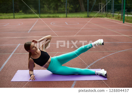 Fit woman exercising outdoors on a yoga mat, performing a side leg lift on a sports court, showcasing strength and determination. Workout concept 128804437