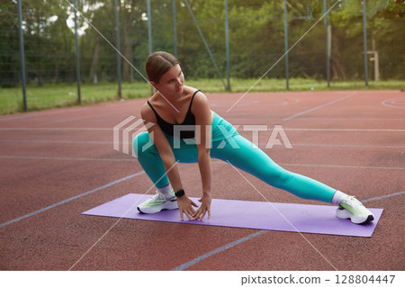 Fit woman stretching on a yoga mat outdoors, showcasing flexibility and strength in a vibrant athletic outfit during fitness routine. Workout concept 128804447