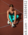 Fit woman tying shoelaces while sitting on a track, showcasing athletic wear and preparing for outdoor exercise session with focus. Workout concept 128804452