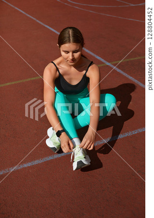 Fit woman tying shoelaces while sitting on a track, showcasing athletic wear and preparing for outdoor exercise session with focus. Workout concept Fit woman tying shoelaces while sitting on a track, showcasing athletic wear and preparing for outdoor exercise session with focus. Workout concept 128804452