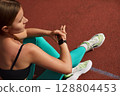 Fit woman checking fitness tracker while sitting on track, wearing stylish athletic gear, showcasing dedication to outdoor exercise and health. Workout concept 128804453
