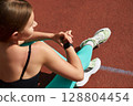 Fit woman checking fitness watch while sitting on track, wearing athletic gear, showcasing dedication to health and outdoor exercise. Workout concept 128804454