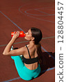 Fit woman in turquoise leggings and black sports top drinking from orange water bottle while sitting on outdoor court, promoting healthy lifestyle. Workout concept 128804457