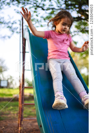 Young girl joyfully sliding down a playground slide in nature 128804603