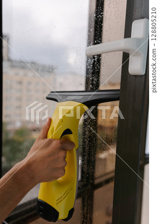 Close-up of male hand using cordless vacuum cleaner for cleaning window, wiping away water drops on rainy day, ensuring spotless and hygienic indoor environment. Concept of household cleanliness. 128805210