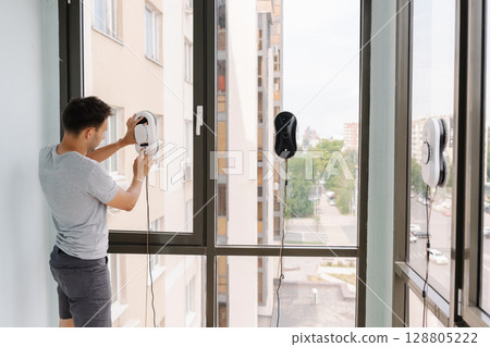 Housekeeper male using advanced robotic device for cleaning large window in modern high-rise, representing innovative window maintenance technology. Handy electronic device for futuristic housekeeping Housekeeper male using advanced robotic device for cleaning large window in modern high-rise, representing innovative window maintenance technology. Handy electronic device for futuristic housekeeping 128805222