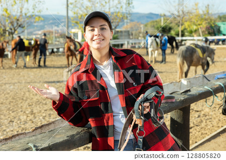 Skilled European young horsewoman in plaid shirt holding harness in hands during work on ranch Skilled European young horsewoman in plaid shirt holding harness in hands during work on ranch 128805820