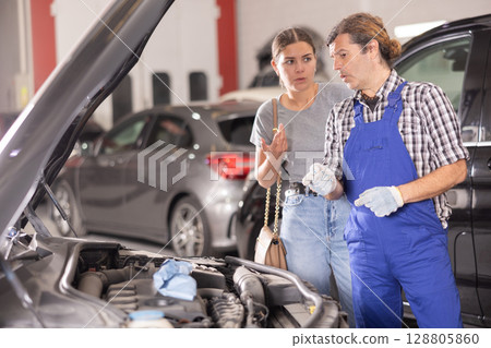 Woman talking to auto mechanic in uniform about repairing his car engine in auto repair shop Woman talking to auto mechanic in uniform about repairing his car engine in auto repair shop 128805860