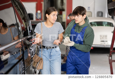 Upset girl owner of car points to scratches on car after an accident to an auto mechanic in car service center Upset girl owner of car points to scratches on car after an accident to an auto mechanic in car service center 128805870