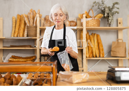 Mature saleswoman putting croissants in plastic bag in bakery Mature saleswoman putting croissants in plastic bag in bakery 128805875